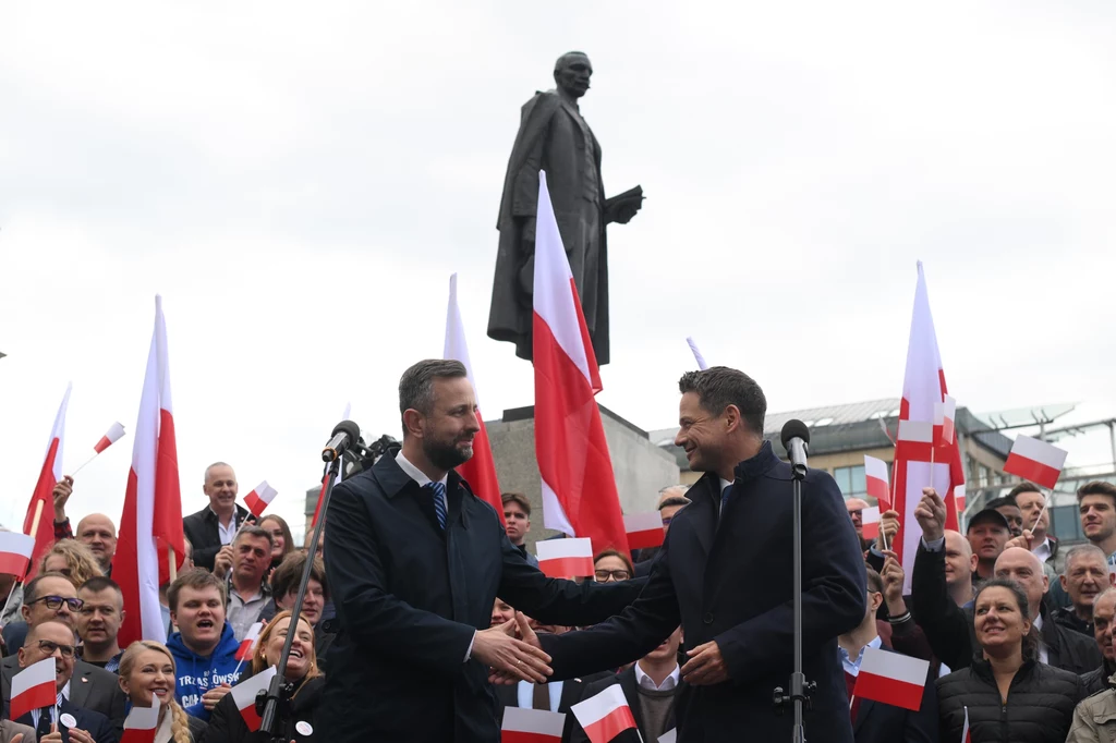 Władysław Kosiniak-Kamysz przekazujący poparcie Rafałowi Trzaskowskiemu przed pomnikiem Witosa w Warszawie Tłum zgromadzonych ludzi z biało-czerwonymi flagami trzymającymi w rękach, dwóch mężczyzn uściskiem dłoni na pierwszym planie, w tle monumentalny pomnik.
