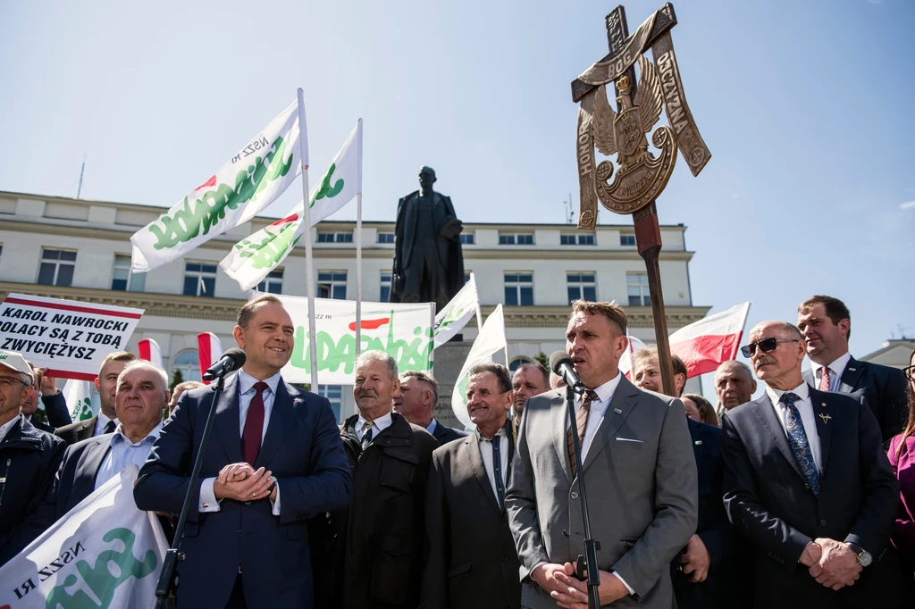Tomasz Obszański, lider NSZZ Rolników Indywidualnych (z prawej) wsparł Karola Nawrockiego w wyborach prezydenckich na konferencji przed pomnikiem Witosa w Warszawie Grupa mężczyzn ubranych w garnitury oraz osoby z flagami i transparentami z napisem 'Solidarność' zgromadzeni przed pomnikiem, wokół unosi się duży drewniany krzyż z orłem, całość na tle budynku.