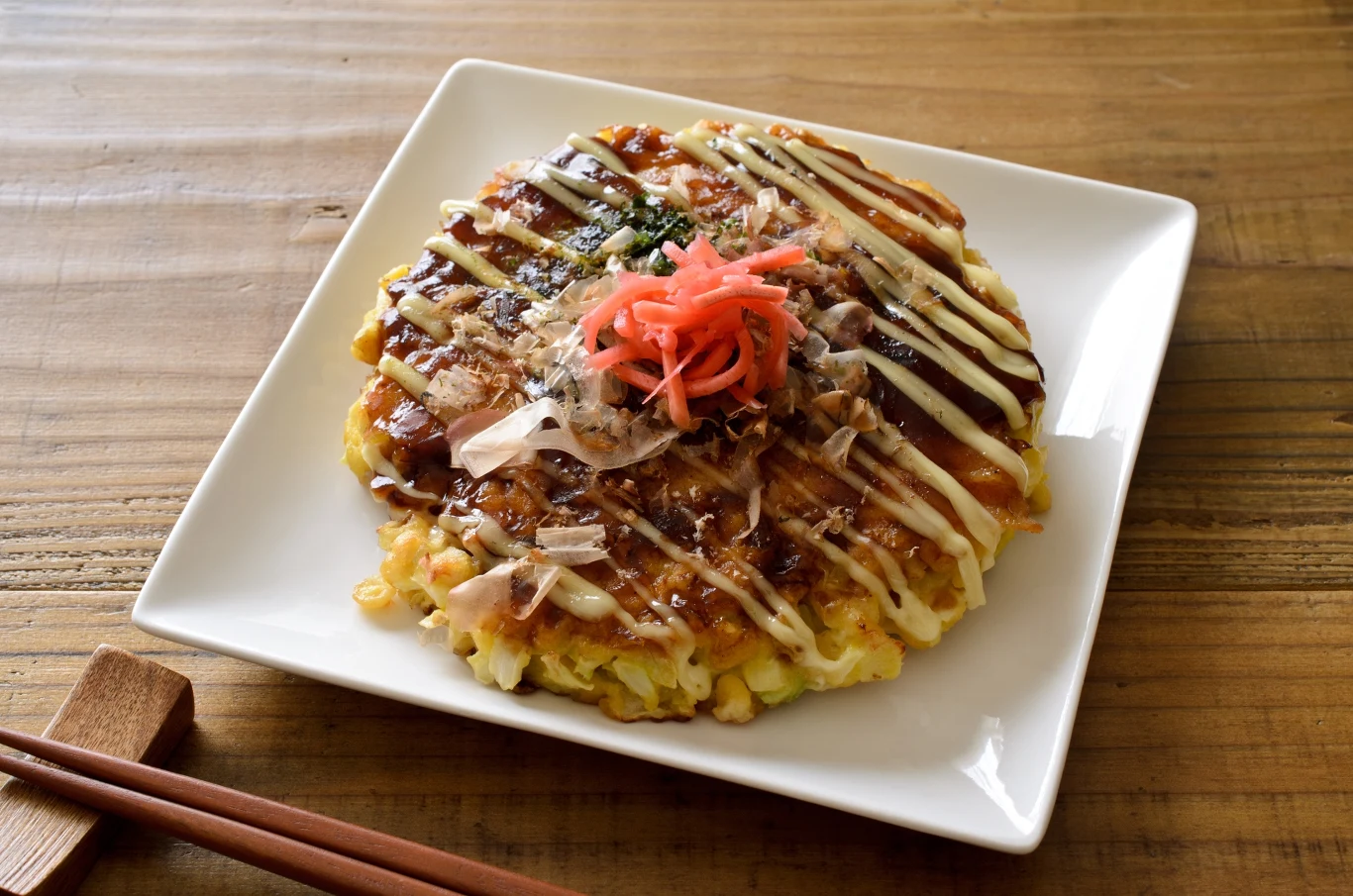 Tradycyjny japoński placek okonomiyaki polany sosem, udekorowany majonezem, katsuobushi oraz czerwoną marynowaną imbirem, podany na kwadratowym talerzu z pałeczkami obok.