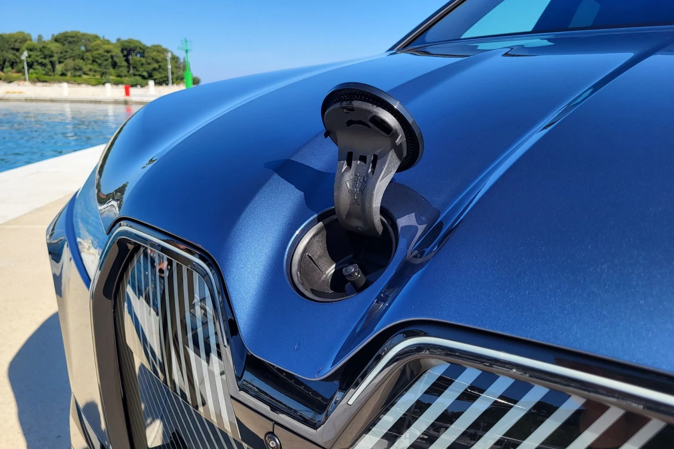 Zbliżenie na przednią maskę niebieskiego samochodu elektrycznego z otwartą klapką portu ładowania, odbijającą światło słoneczne, w tle fragment nadbrzeża i drzewa.