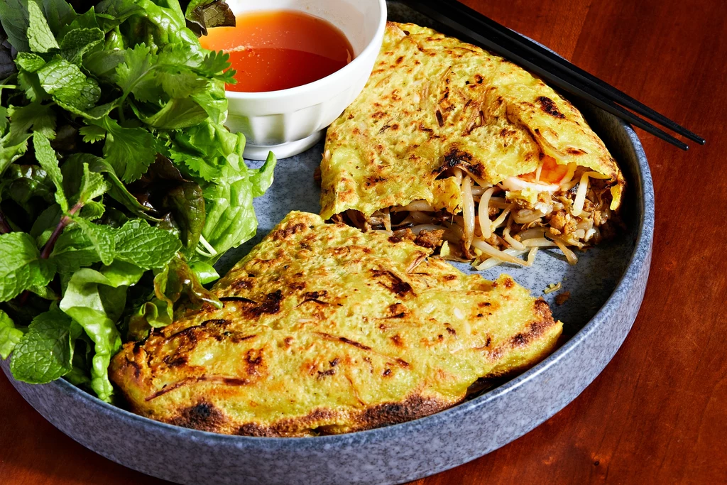 Bánh xèo jest szczególnie popularne w południowym Wietnamie i często pojawia się na ulicznych straganach oraz domowych przyjęciach Żółty omlet podany na pół, wypełniony kiełkami fasoli i innymi warzywami, obok świeże zioła i miseczka z sosem, całość ułożona na okrągłym talerzu.