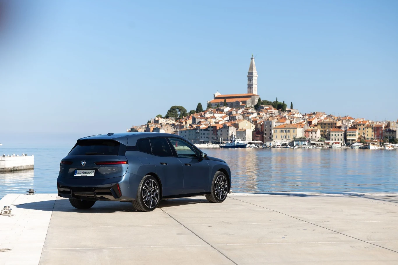 Nowoczesny czarny samochód zaparkowany na betonowym nabrzeżu, z widokiem na malownicze nadmorskie miasteczko ze starą zabudową oraz charakterystyczną wieżą kościelną w tle.