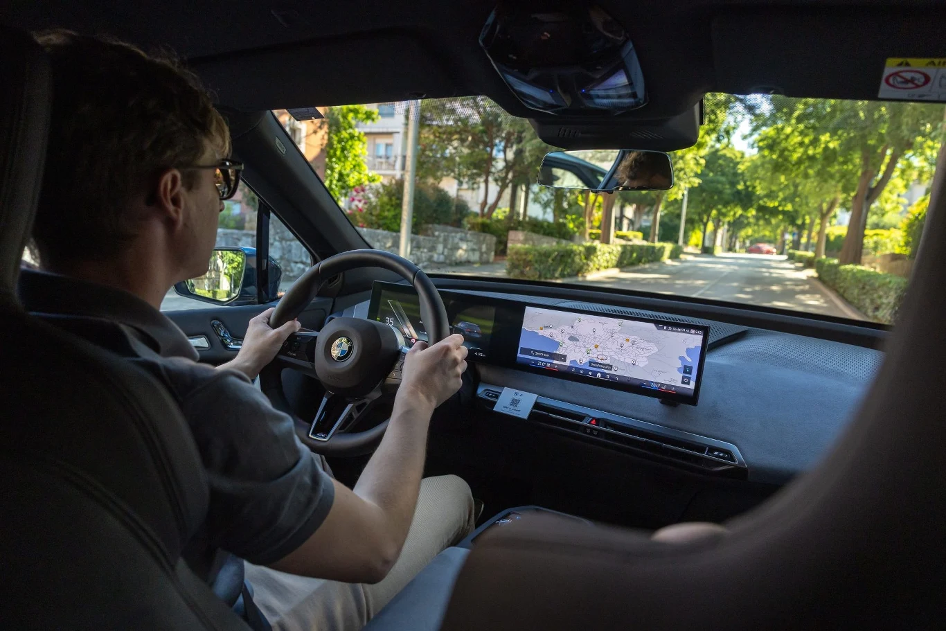 Wnętrze nowoczesnego samochodu podczas jazdy po miejskiej ulicy, kierowca trzyma kierownicę, na konsoli widoczny duży, panoramiczny ekran nawigacji, przez szyby widać zieleniejącą okolicę i zabudowę mieszkalną.