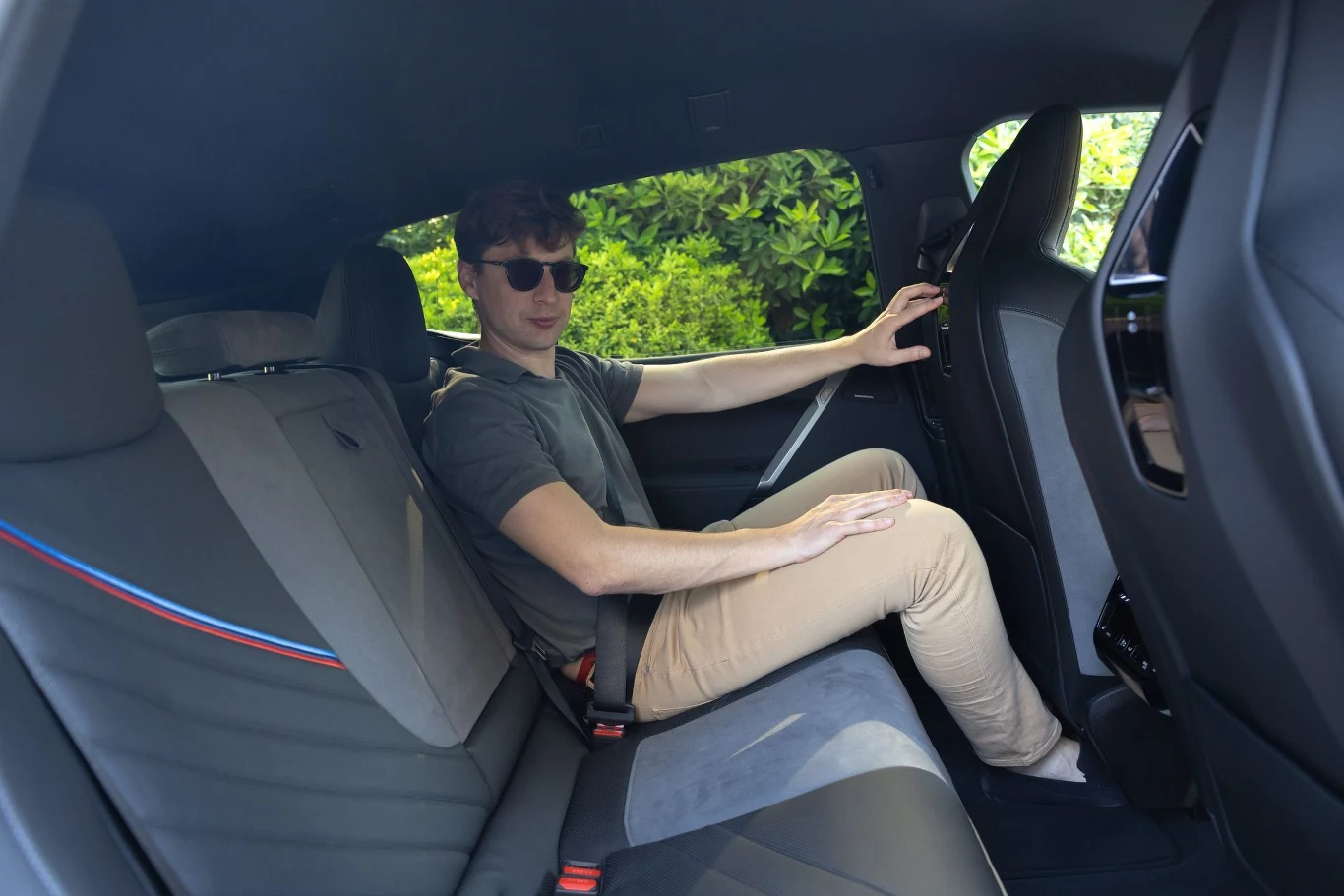 Mężczyzna w okularach przeciwsłonecznych siedzi na tylnym siedzeniu samochodu, opierając jedną rękę na oparciu przedniego fotela, wnętrze auta przestronne, w tle widoczne zielone krzewy za oknem.