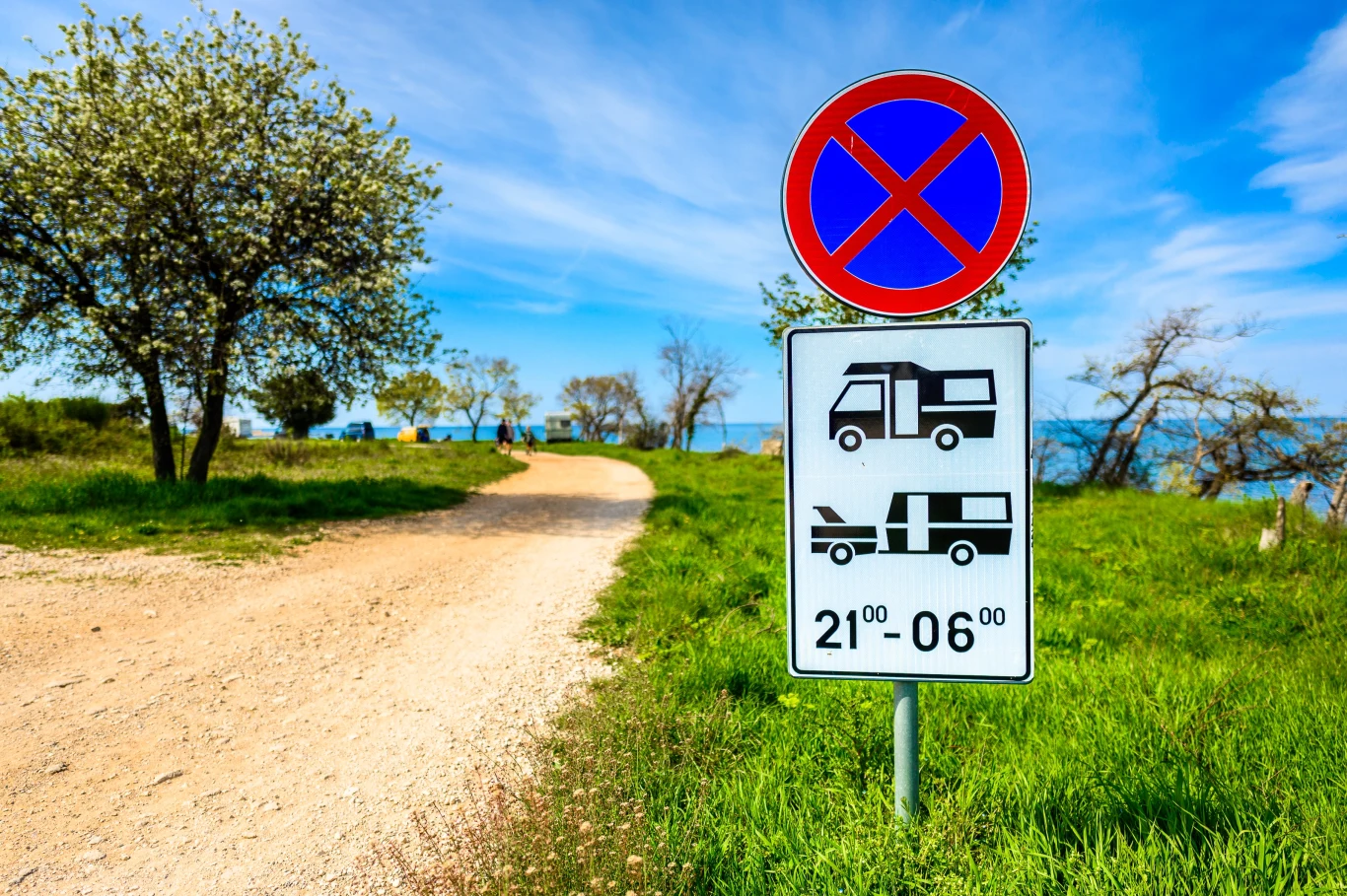 Wiele państw w pewnym stopniu reguluje ruch kamperów. Na zdjęciu jeden z zakazów w Chorwacji Droga gruntowa prowadząca w stronę wody, po bokach zielona trawa i drzewa, przy drodze stoi znak zakazu parkowania oraz informacja o zakazie parkowania kamperów i przyczep campingowych w godzinach nocnych.
