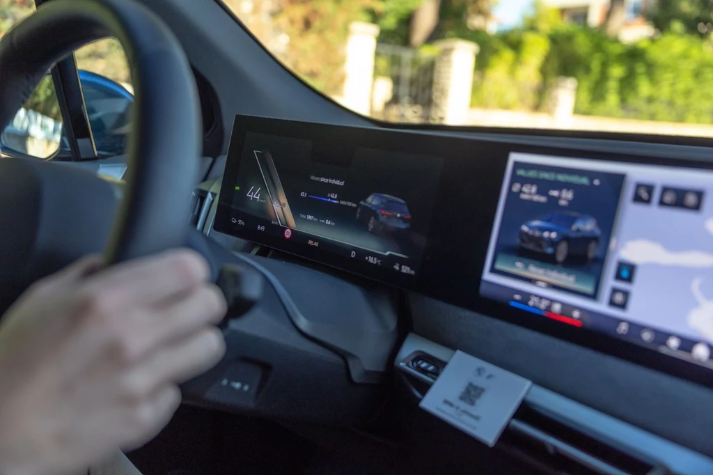 Ekran przed kierowcą łączy się płynnie z wyświetlaczem multimediów, podobnie jak to jest w wielu innych modelach BMW Wnętrze nowoczesnego samochodu z cyfrowymi ekranami na desce rozdzielczej, na których prezentowane są informacje dotyczące jazdy oraz multimediów, widoczna lewa ręka kierowcy na kierownicy.