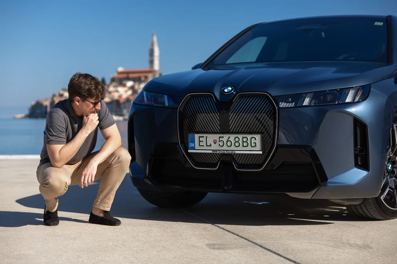 Mężczyzna w okularach przeciwsłonecznych w przyklęku obok nowoczesnego samochodu marki BMW, z widocznym grillem i tablicą rejestracyjną, w tle nadmorskie miasto z wyraźnie widoczną wieżą kościelną.
