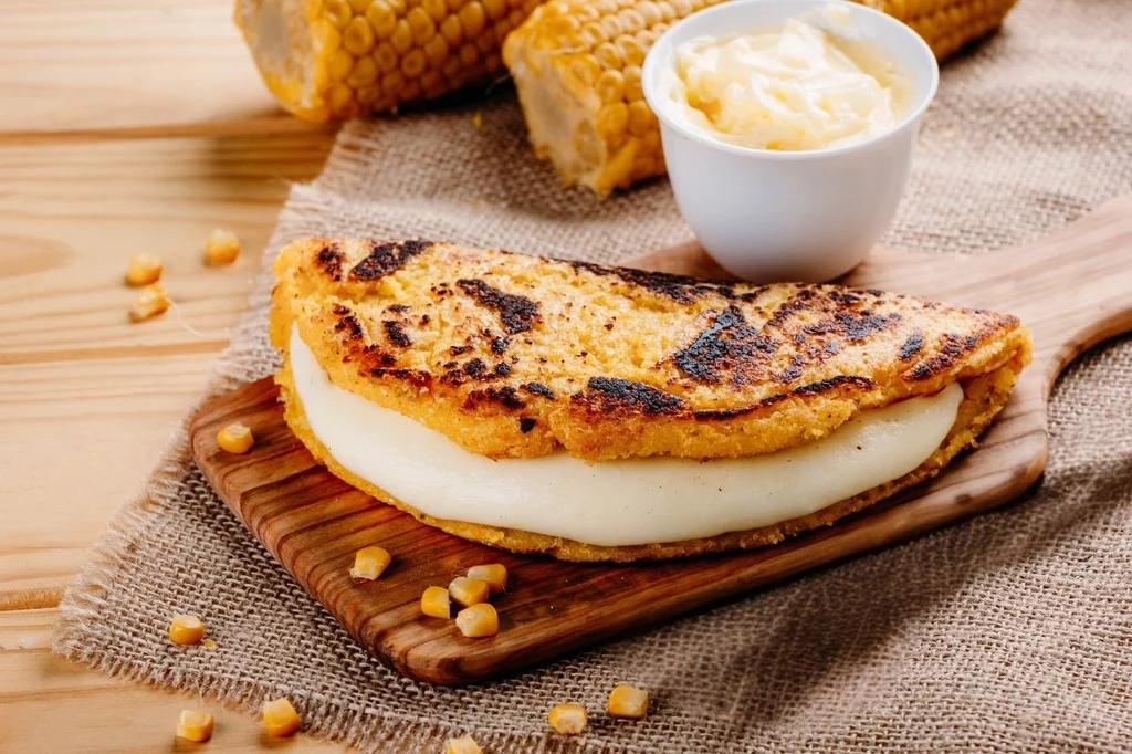 Cachapas to wenezuelski przysmak, który króluje na tamtejszych ulicach Grillowana plackowata tortilla z serem w środku na drewnianej desce, obok znajduje się miseczka z masłem oraz kilka ziaren kukurydzy i kawałki kolby kukurydzy w tle.