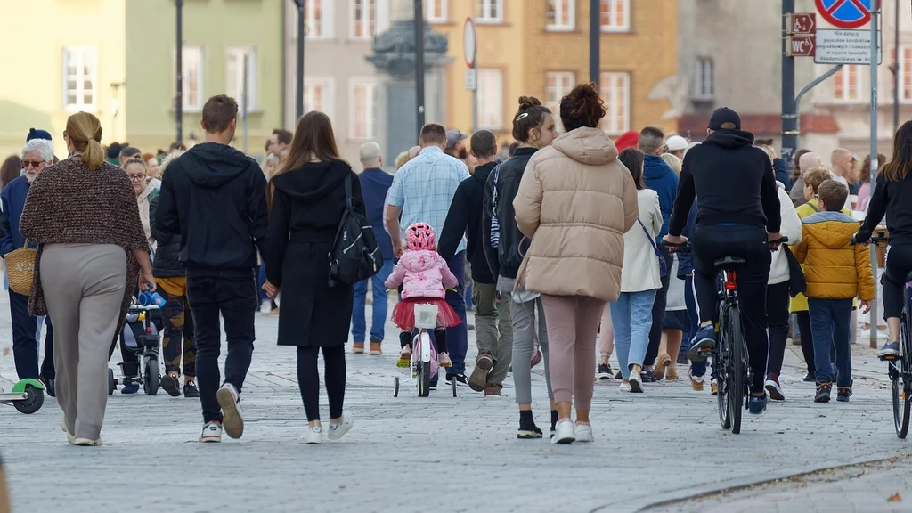 Fot. pixabay.com/ Surprising_Media Tłum ludzi spacerujących po miejskim deptaku, wśród nich dzieci na rowerkach i osoby na hulajnogach oraz rowerach, widoczne różnorodne ubrania listopadowe, w tle kolorowe kamienice.