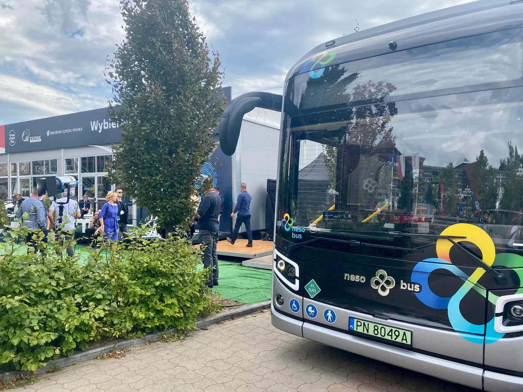 XXXI Forum Ekonomicznego w Karpaczu Nowoczesny autobus wystawiony podczas targów lub wydarzenia branżowego, wokół którego gromadzą się odwiedzający i zwiedzający, po lewej stronie widoczne krzewy i drzewa, w tle znajdują się stoiska wystawiennicze.