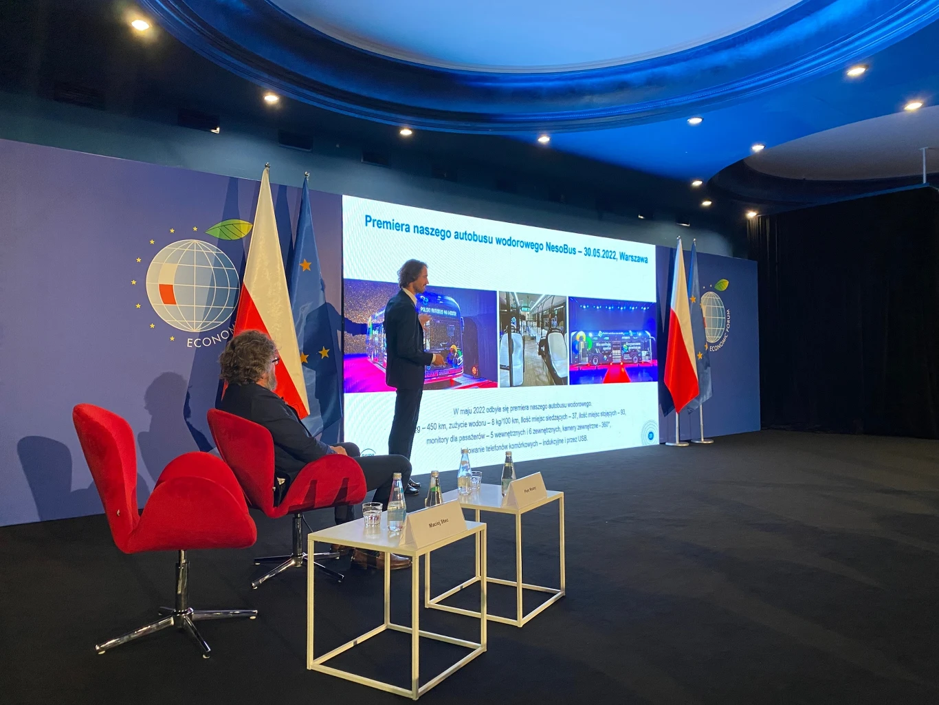 Dwóch mężczyzn uczestniczących w oficjalnej prezentacji na tle dużego ekranu multimedialnego, na którym widoczne są zdjęcia i tekst o premierze autobusu wodorowego, po bokach flagi Polski, Unii Europejskiej oraz czerwone krzesła i stoły z wodą.