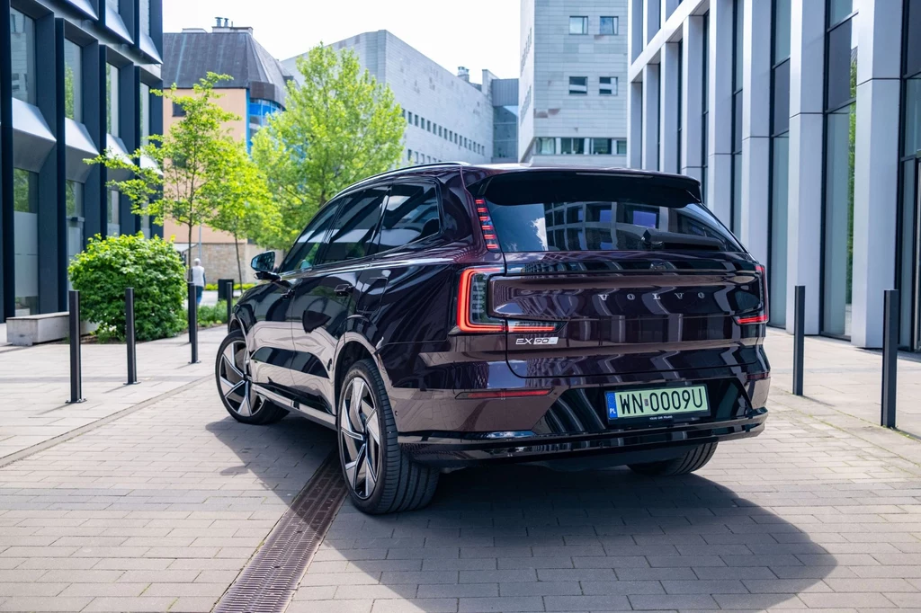 Tylne lampy mają charakterystyczny, pionowy kształt Nowoczesny, ciemnofioletowy SUV marki EXEED TXL stoi zaparkowany między miejskimi budynkami, otoczony szklaną architekturą i zielenią.
