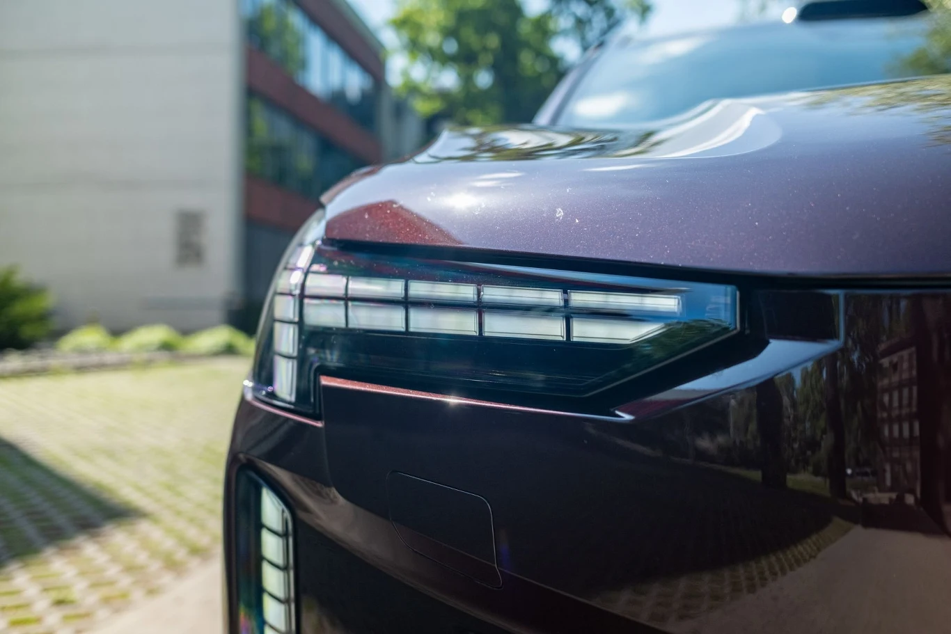 Przedni reflektor nowoczesnego samochodu w ciemnym kolorze, odbijający światło słoneczne, na tle budynków i zieleni w miejskim otoczeniu.