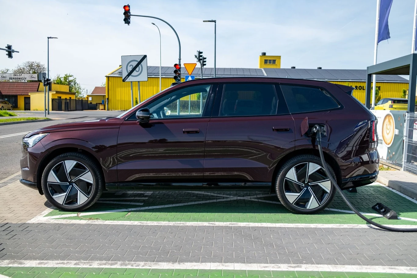 SUV o ciemnej karoserii podłączony do stacji ładowania pojazdów elektrycznych na tle nowoczesnych budynków i znaków drogowych, z wyraźnie widocznym przewodem ładującym.