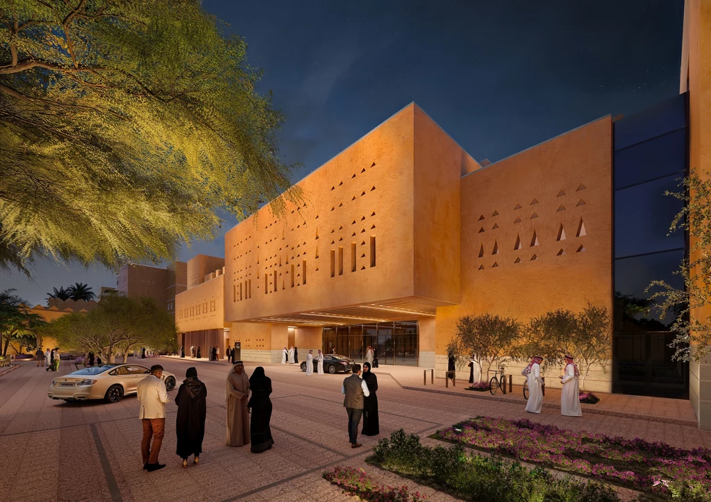 Nowoczesna architektura z błota w sercu Arabii Saudyjskiej Nowoczesny budynek wykonany w stylu arabskim, w ciepłych, piaskowych barwach oświetlony sztucznym światłem, z charakterystycznymi ozdobnymi otworami w elewacji, otoczony roślinnością, po chodniku spacerują osoby ubrane w tradycyjne stroje.