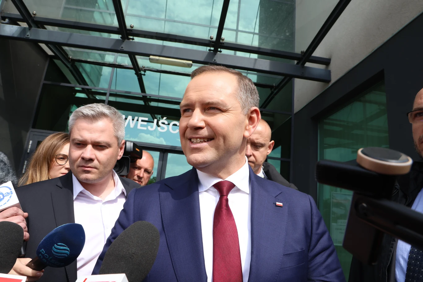 Karol Nawrocki tuż po spotkaniu ze Sławomirem Mentzenem w jego kancelarii w Toruniu Mężczyzna w garniturze i czerwonym krawacie stoi na zewnątrz budynku z napisem 'WEJŚCIE', otoczony przez dziennikarzy z mikrofonami, uśmiecha się, za nim kilka innych osób.