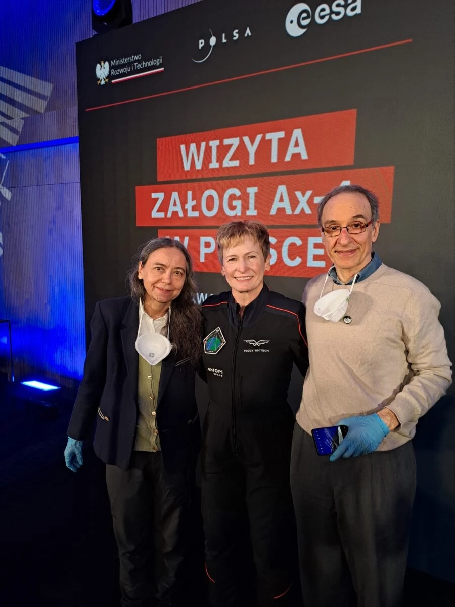 Przedstawiciele zespołu Yeast TardigradeGene z Uniwersytetu Szczecińskiego: Ewa Szuszkiewicz i Franco Ferrari wraz z dowódcą misji Ax-4 – Peggy Whitson. Trójka dorosłych osób pozuje na tle czarnego baneru z napisami dotyczącymi wizyty załogi Axiom w Polsce, dwie osoby ubrane są w garnitury, jedna w skafander kosmiczny, a wszyscy wyglądają na zadowolonych.