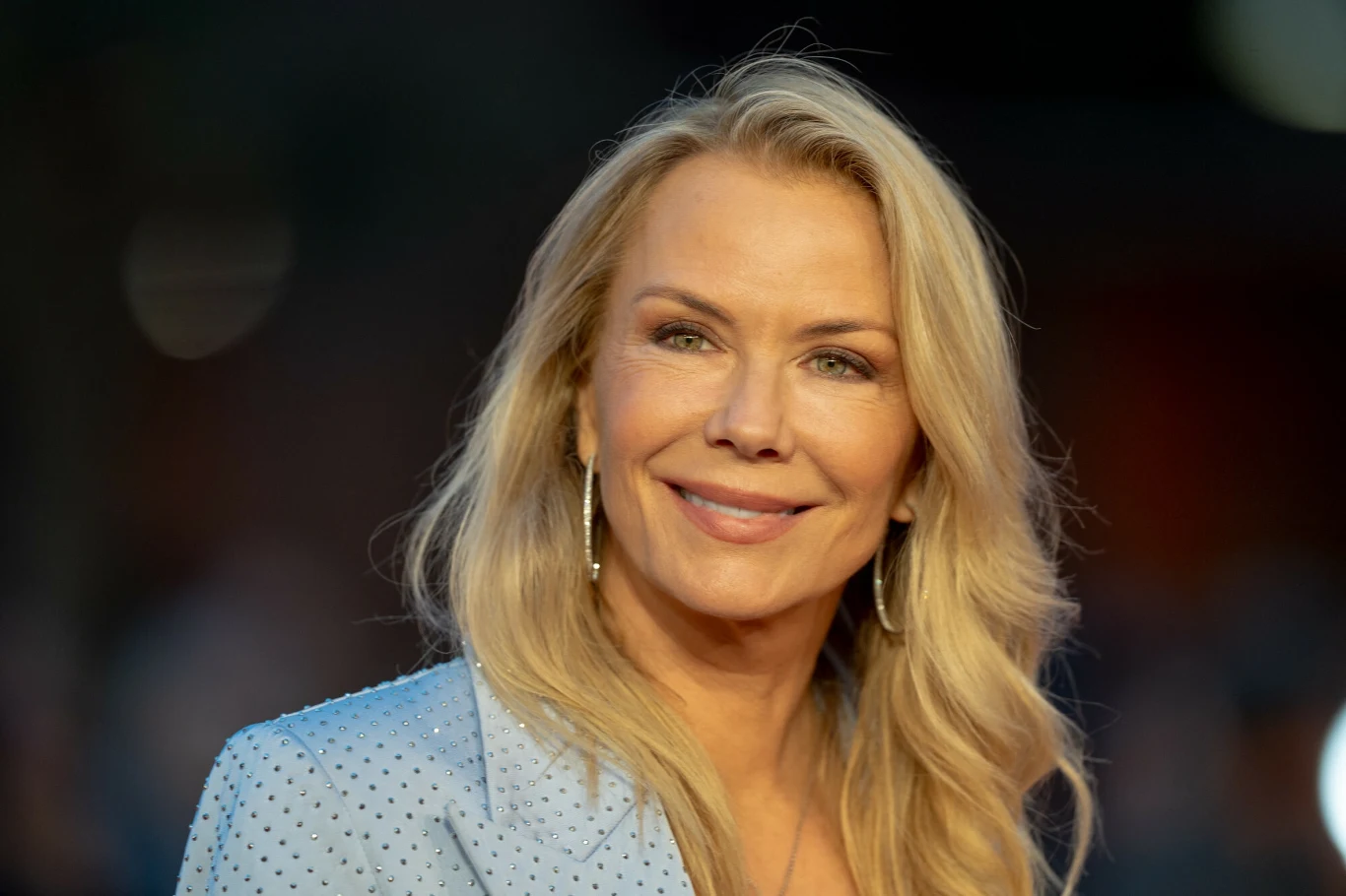Uśmiechnięta kobieta o blond włosach ubrana w jasnoniebieską marynarkę, stojąca na tle rozmytego, ciemnego tła, patrząca bezpośrednio w obiektyw.