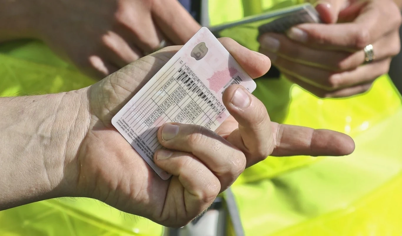 Kody w prawie jazdy nakładają na kierowcę konkretne oznaczenia Ręka trzymająca plastikowy dokument prawdopodobnie prawo jazdy, w tle fragment żółtej odblaskowej kamizelki oraz druga osoba trzymająca kartę.