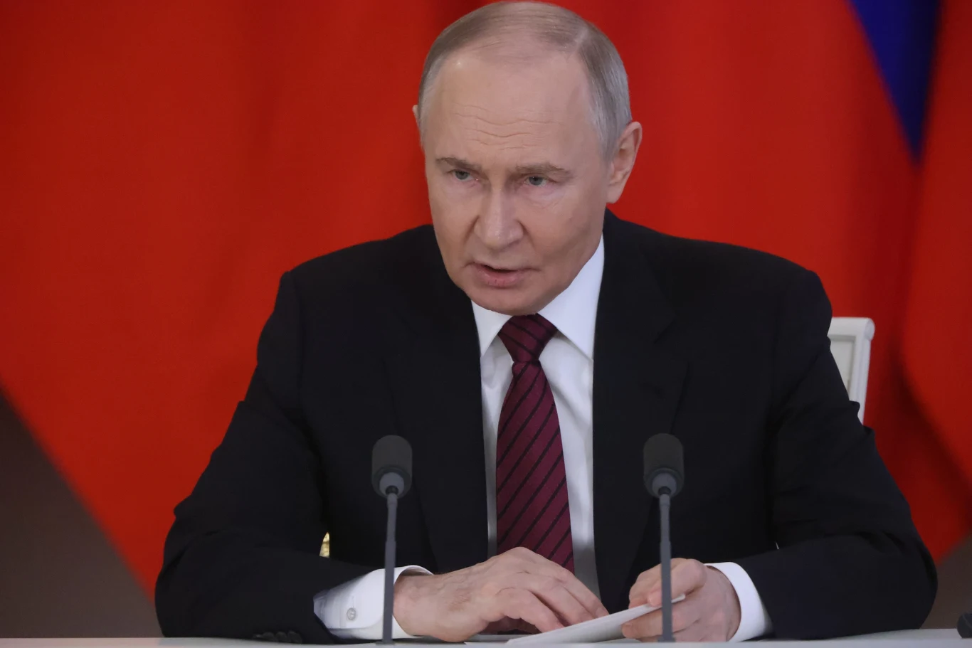 Władimir Putin wprowadza strefę buforową wzdłuż granic Rosji Mężczyzna w średnim wieku w garniturze i krawacie siedzi za stołem z dwoma mikrofonami, w tle rozpoznawalne czerwone tło z fragmentem niebieskiego koloru, sugerujące obecność flagi.