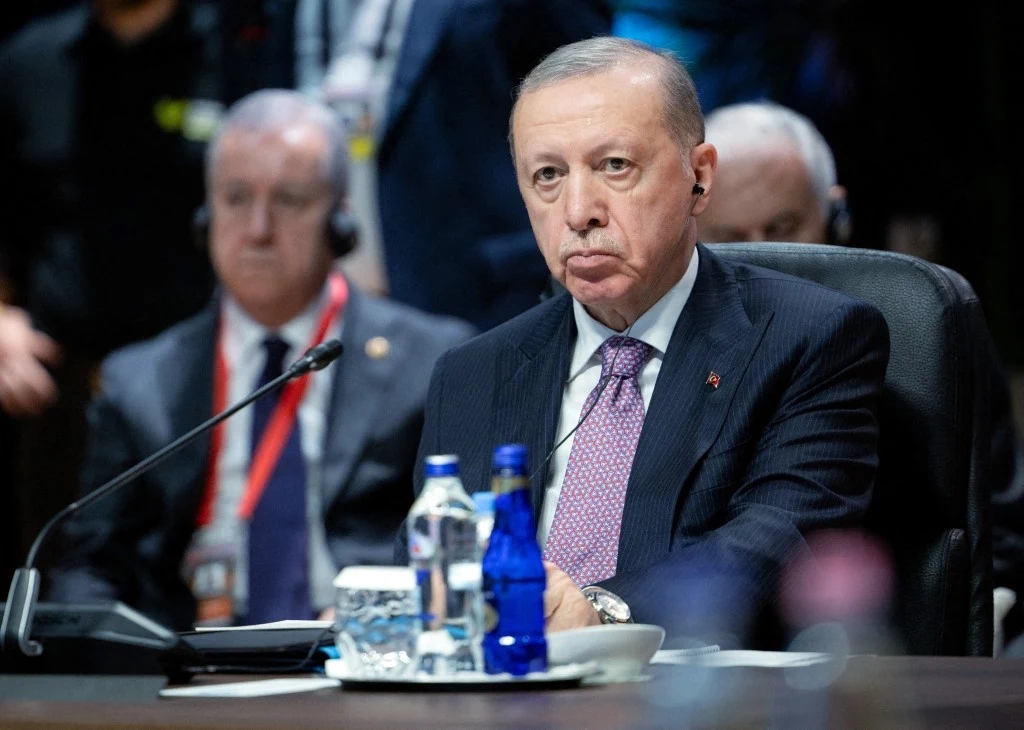 Starszy mężczyzna w garniturze i krawacie, siedzący przy stole konferencyjnym, słuchający przez słuchawkę, przed nim butelki wody i filiżanka, w tle inni uczestnicy spotkania.