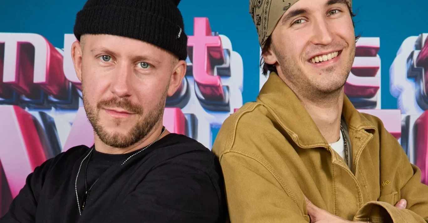 Alien x Majtis - zwycięzcy "Must Be The Music" Dwóch mężczyzn stojących tyłem do siebie, jeden ubrany w czarną czapkę i czarną koszulkę, drugi w jasną bandanę i brązową kurtkę, obaj uśmiechnięci, w tle kolorowe napisy.