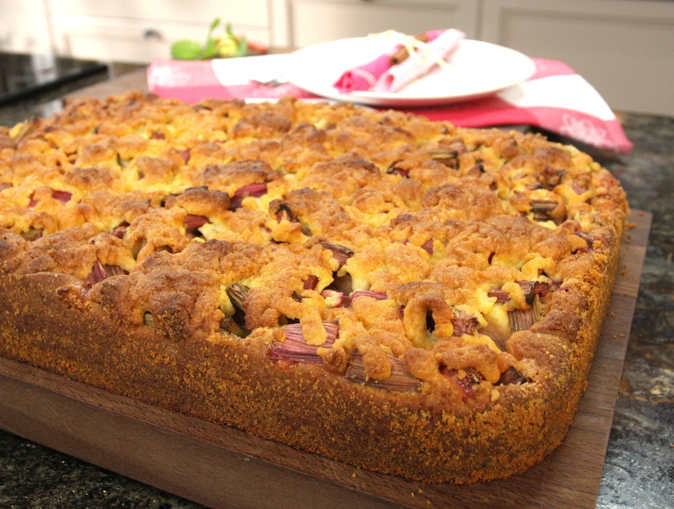 Warto wykorzystać sezon na rabarbar Duże, złociste ciasto drożdżowe z kruszonką i kawałkami rabarbaru, podane na drewnianej desce, w tle biały talerz z różowym serwetnikiem.