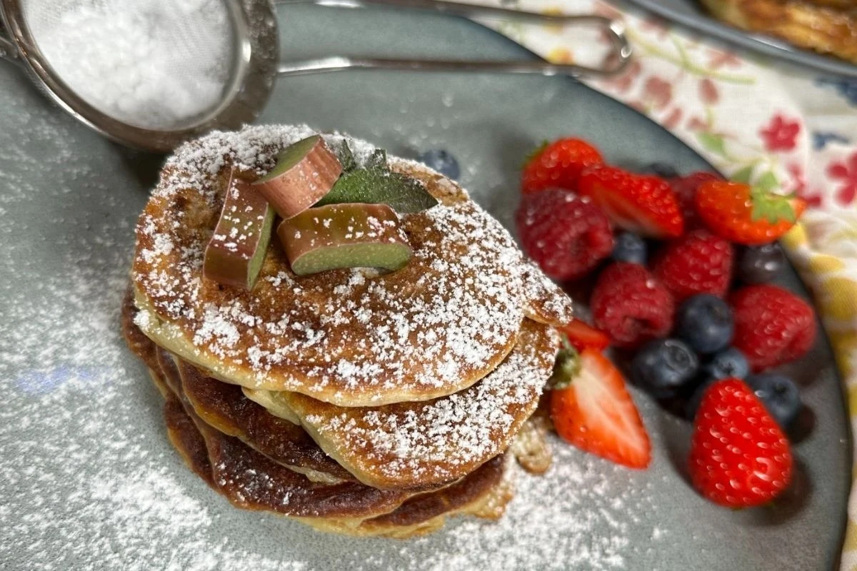 Racuchy z rabarbarem to świetna propozycja dla najmłodszych Stos naleśników posypanych cukrem pudrem, udekorowanych kawałkami rabarbaru i listkami mięty, obok świeże owoce: truskawki, maliny, borówki i jeżyny.