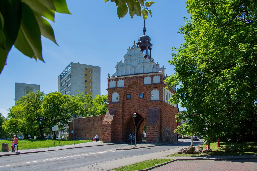 Prace trwają przy ul. Chrobrego, gdzie znajduje się Brama Wałowa. Historyczna brama miejska w otoczeniu zielonych drzew, za nią widoczne wysokie bloki mieszkalne, jasne niebo i spokojna ulica biegnąca pod bramą.