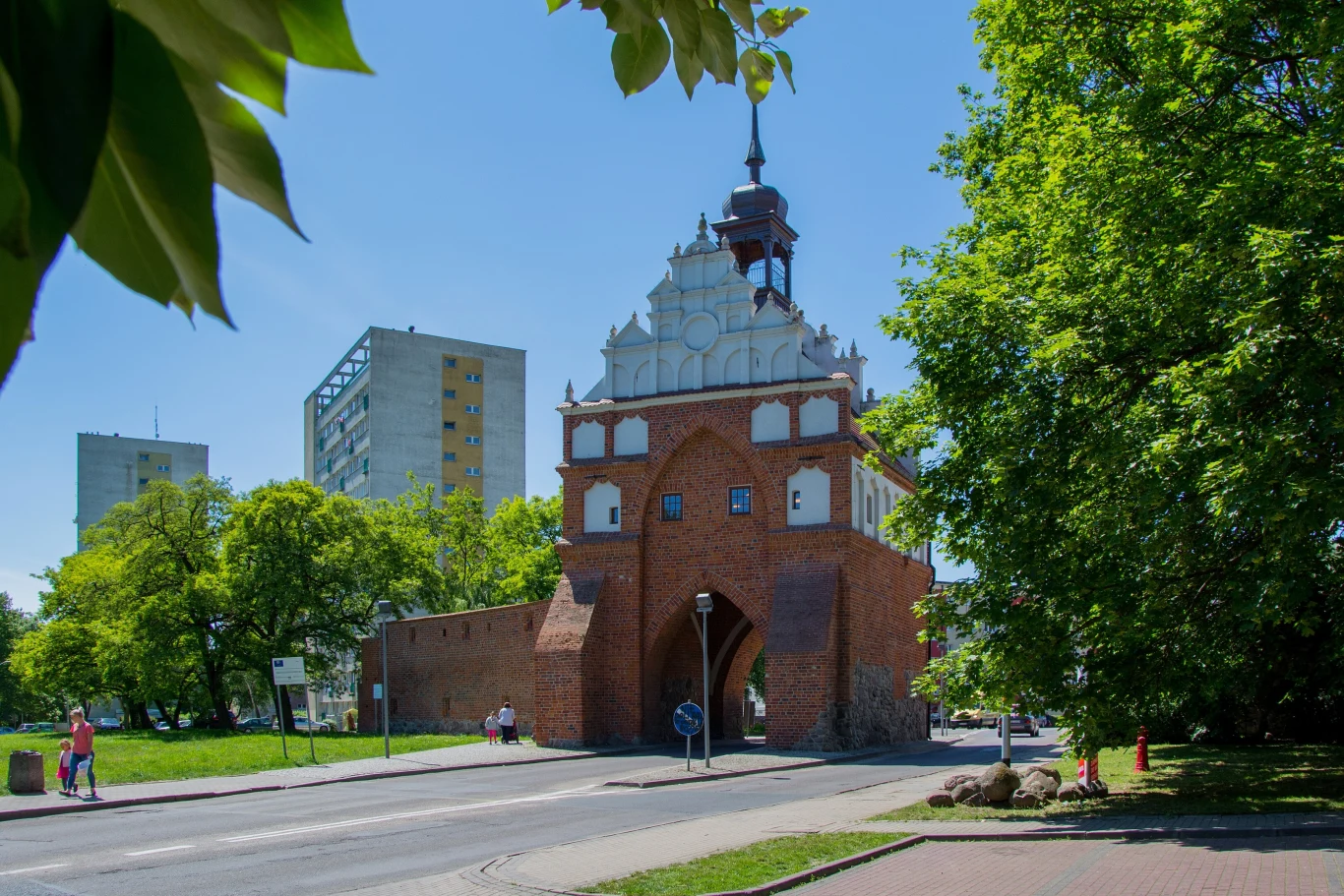Prace trwają przy ul. Chrobrego, gdzie znajduje się Brama Wałowa. Historyczna brama miejska w otoczeniu zielonych drzew, za nią widoczne wysokie bloki mieszkalne, jasne niebo i spokojna ulica biegnąca pod bramą.
