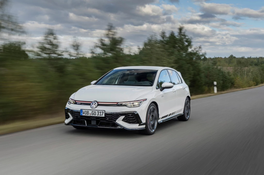 Szukacie VW Golfa w sportowej wersji? To idealny moment na zakup w niższych cenach. Biały samochód osobowy marki Volkswagen Golf poruszający się po pustej, asfaltowej drodze obok lasu, w tle niebo z chmurami.
