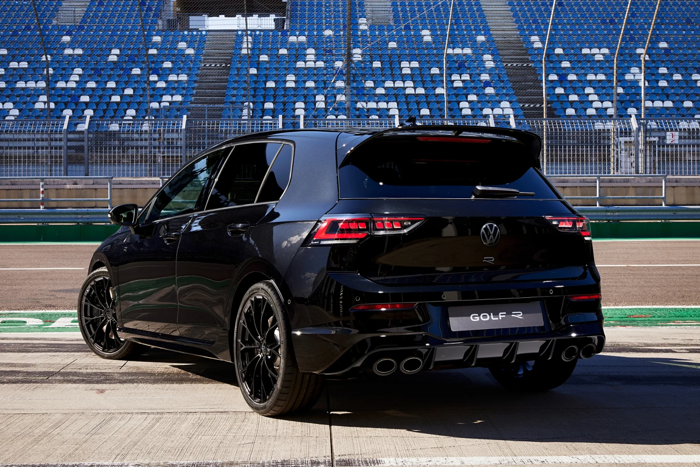 Czarny samochód Volkswagen Golf R zaparkowany na torze wyścigowym, w tle widoczne puste trybuny niebiesko-białe i zabezpieczenia toru.
