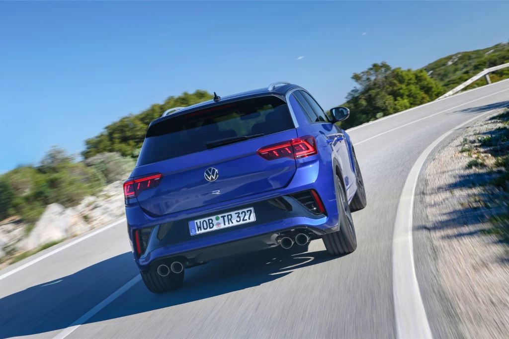 T-Roc R pod maską posiada silnik 2.0 TSI o mocy 300 KM. Niebieski samochód Volkswagen jadący po krętej, asfaltowej drodze, otoczonej zielenią i niskimi wzgórzami. Auto widziane od tyłu, dynamiczna scena w ruchu.