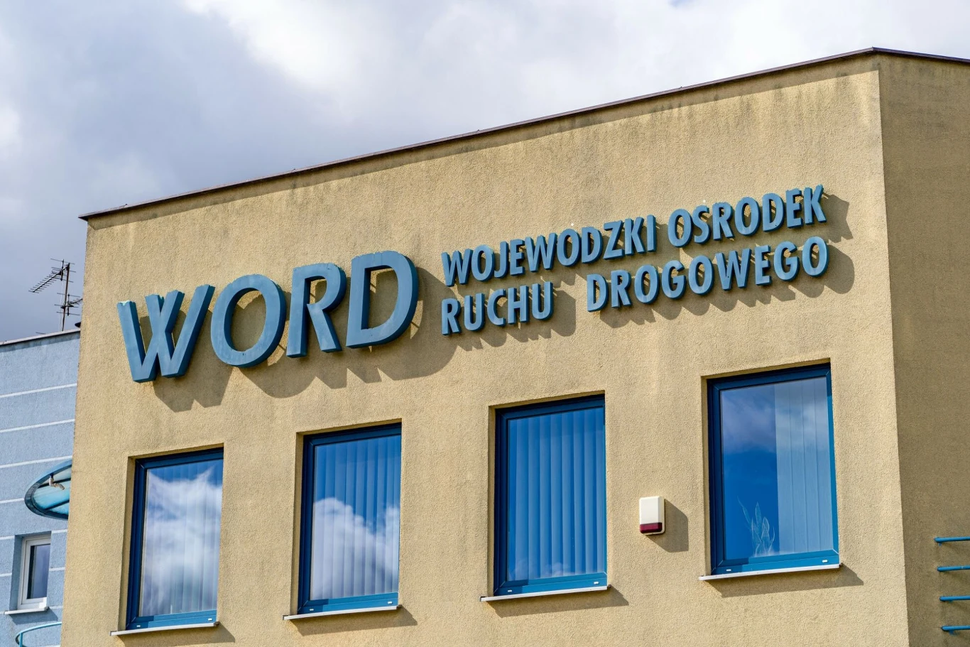 Budynek z napisem WORD Wojewódzki Ośrodek Ruchu Drogowego, trzy niebieskie okna z odbiciem nieba, jasna elewacja podkreślająca charakter urzędowy obiektu.