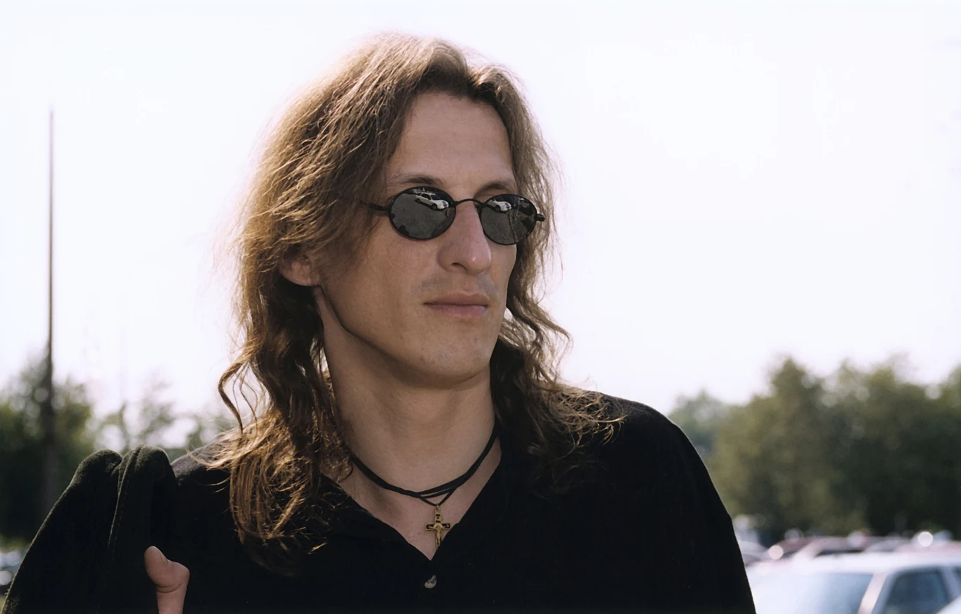 Jacek Dewódzki był następcą Ryśka Riedla w grupie Dżem Mężczyzna o długich włosach i w okrągłych okularach przeciwsłonecznych, ubrany na czarno, stoi na zewnątrz w słoneczny dzień, w tle widoczne drzewa i rozmyte sylwetki samochodów.