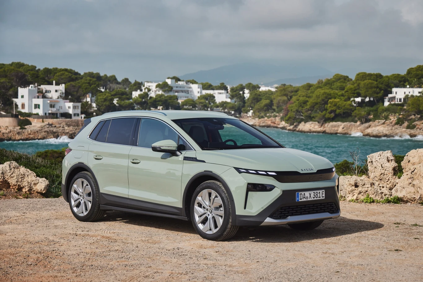 Jasnozielony samochód elektryczny SUV stoi na tle wybrzeża z morzem, wokół roślinność śródziemnomorska i białe domy, w oddali zalesione wzgórza.