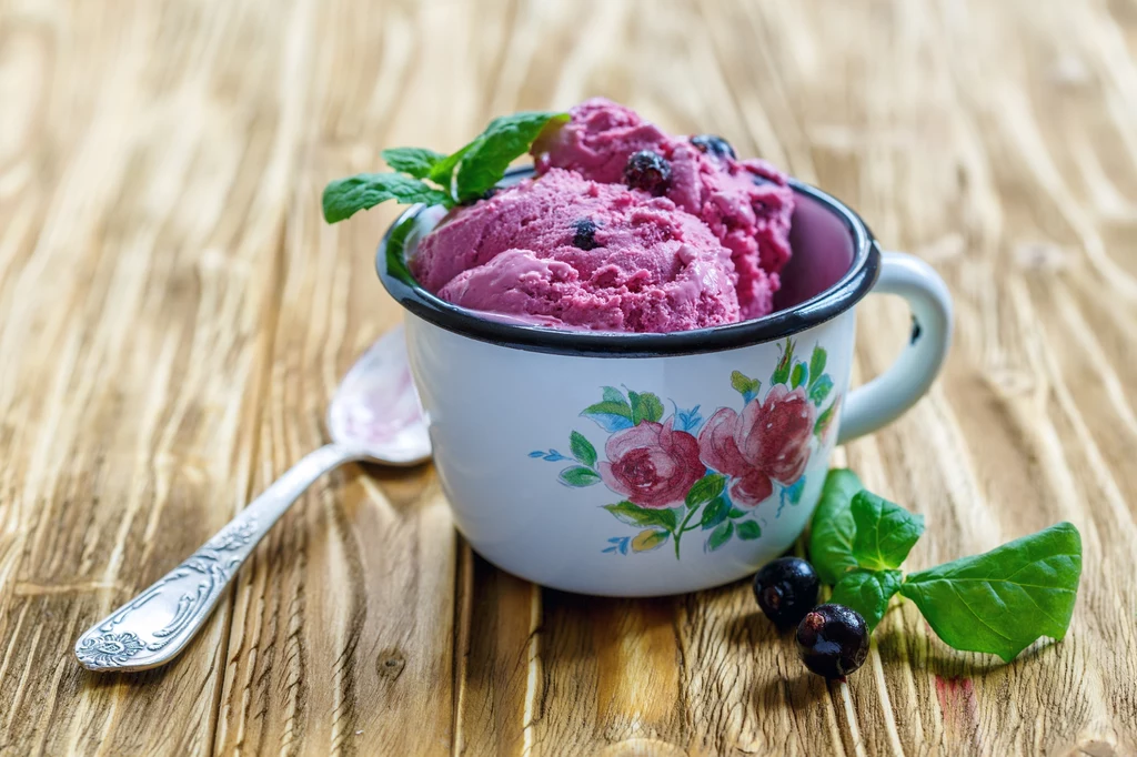 Lody z serka wiejskiego są pełne białka Porcja fioletowych lodów z czarną porzeczką, podana w białym emaliowanym kubku z kwiatowym wzorem, ozdobiona listkiem mięty, na drewnianym stole, obok metalowej łyżki.