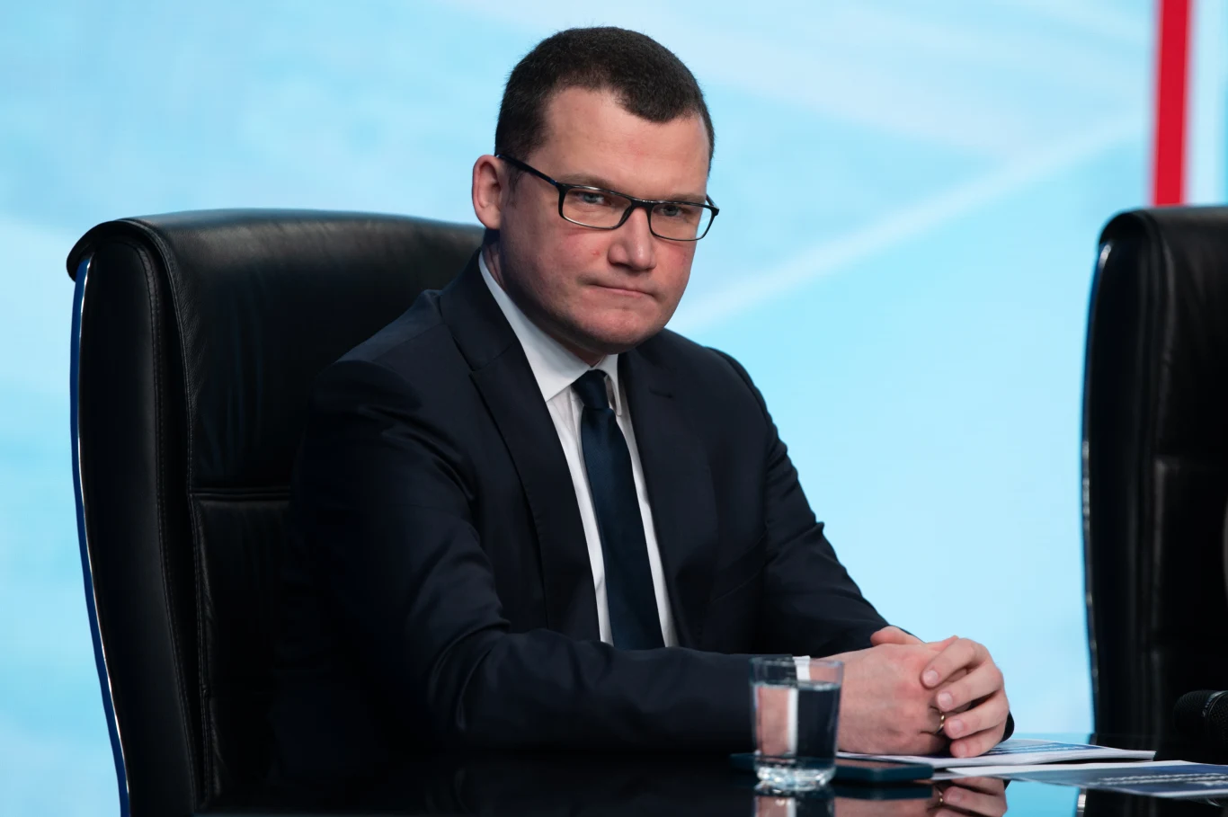 Paweł Szefernaker Mężczyzna w garniturze siedzący przy stole konferencyjnym, z poważnym wyrazem twarzy, przed nim szklanka wody, w tle jasnoniebieskie tło.