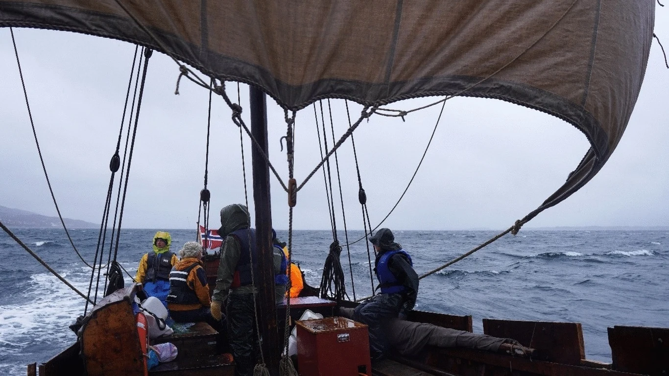 Grupa osób na tradycyjnej łodzi żaglowej płynącej po wzburzonym morzu, pochmurne niebo i widoczne fale podkreślają surowe warunki rejsu.