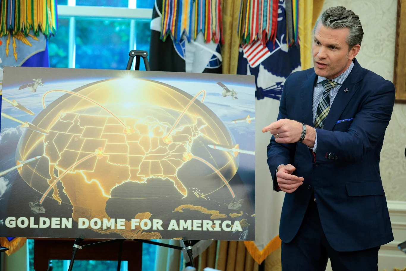 Mężczyzna w garniturze stoi obok planszy z ilustracją półkuli ziemskiej, na której przedstawiono mapę USA oraz trajektorie ruchu rakiet lub pocisków. Plansza podpisana jest hasłem GOLDEN DOME FOR AMERICA, w tle widoczne są flagi i zasłony.