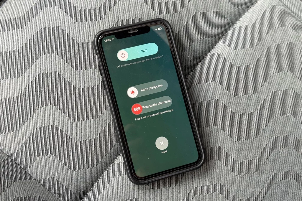 iPhone sam zadzwoni na 112, jeśli ustawimy mu tę możliwość. Smartfon w czarnej obudowie leżący na tapicerce w szaro-białe zygzaki, ekran wyświetla opcje alarmowe takie jak karta medyczna, połączenie alarmowe SOS oraz suwak do wyłączenia urządzenia.