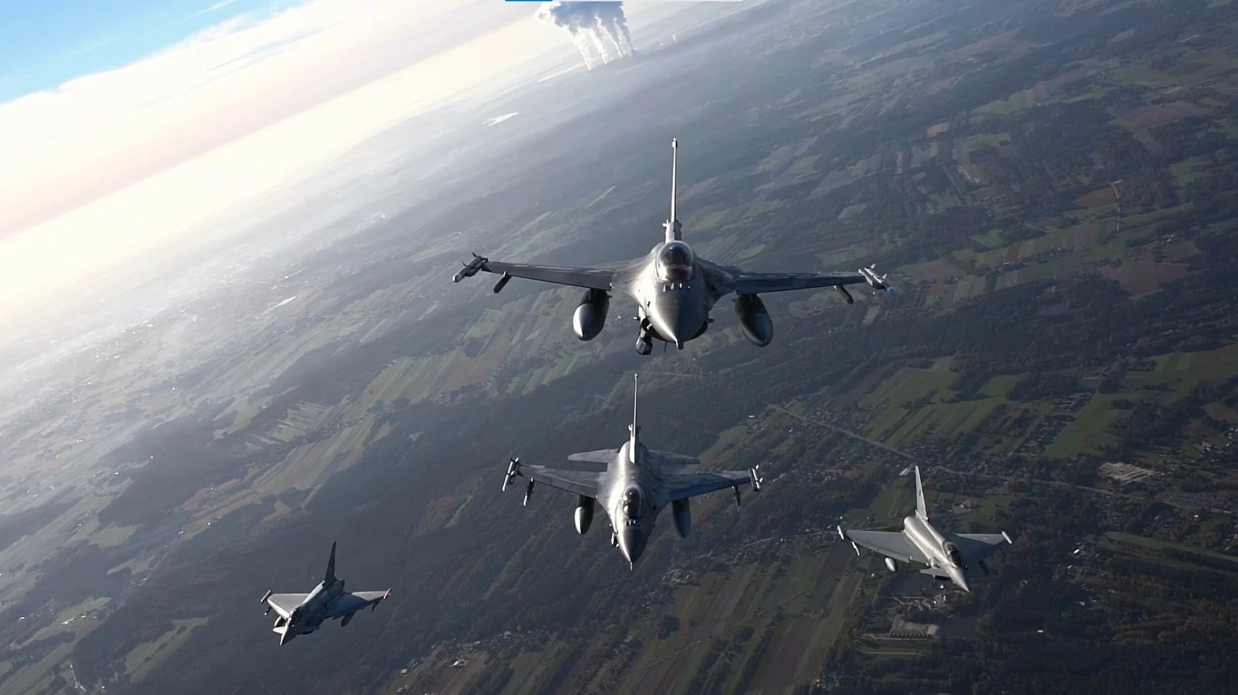 Polskie myśliwce brały udział w akcji NATO (zdjęcie ilustracyjne) Formacja czterech nowoczesnych myśliwców wojskowych lecących nad zielonym krajobrazem z polami i lasami, pod wysokim niebem z lekkim zachmurzeniem.