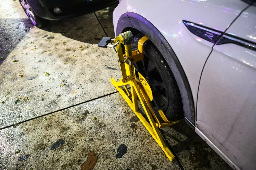 W przypadku nieprawidłowego zaparkowania, kierowca otrzyma mandat. Biała osobówka z blokadą na kole zamontowaną przez służby miejskie, auto stoi na ulicy obok innego pojazdu, na mokrym chodniku widać ślady i zabrudzenia.
