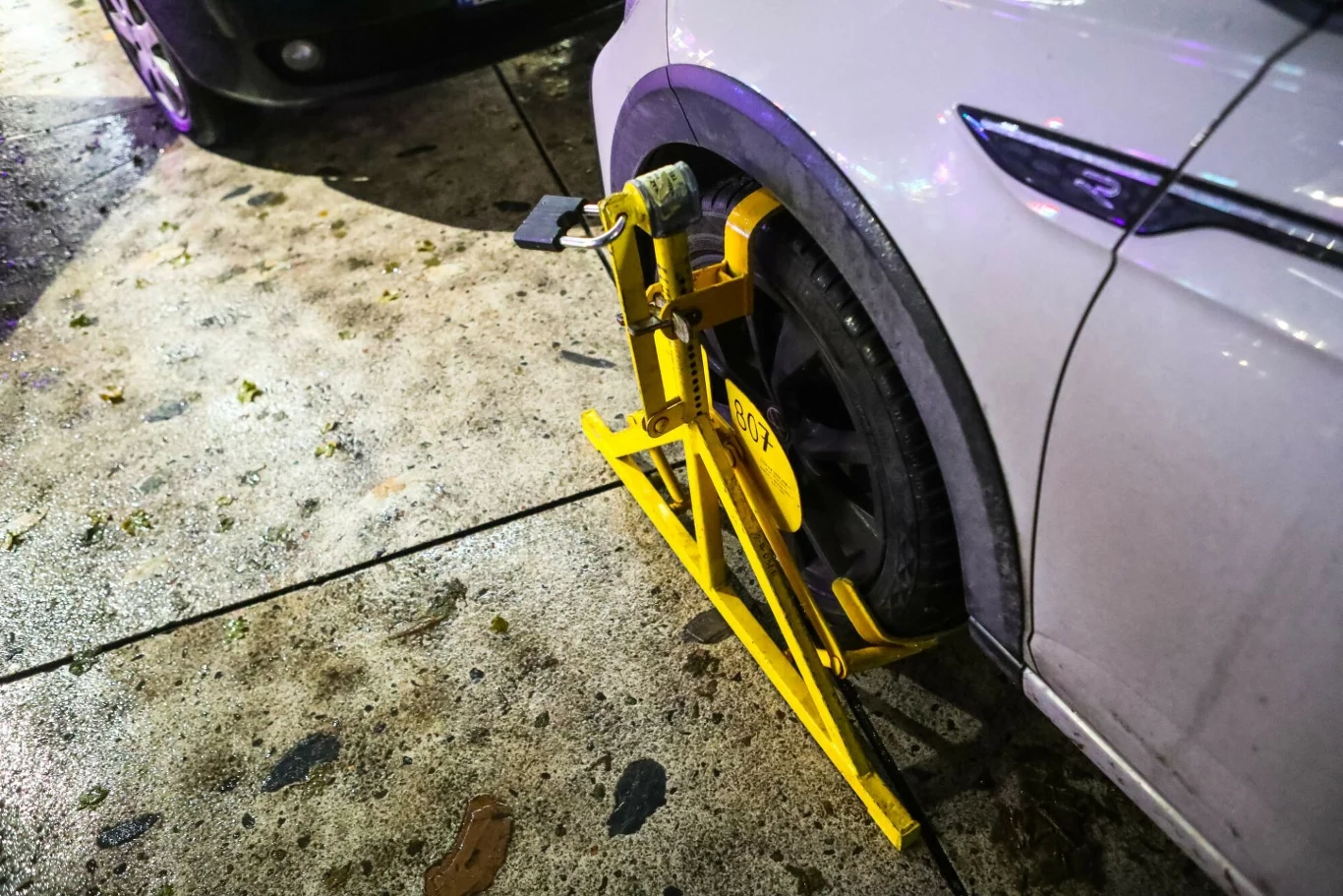 Biała osobówka z blokadą na kole zamontowaną przez służby miejskie, auto stoi na ulicy obok innego pojazdu, na mokrym chodniku widać ślady i zabrudzenia.