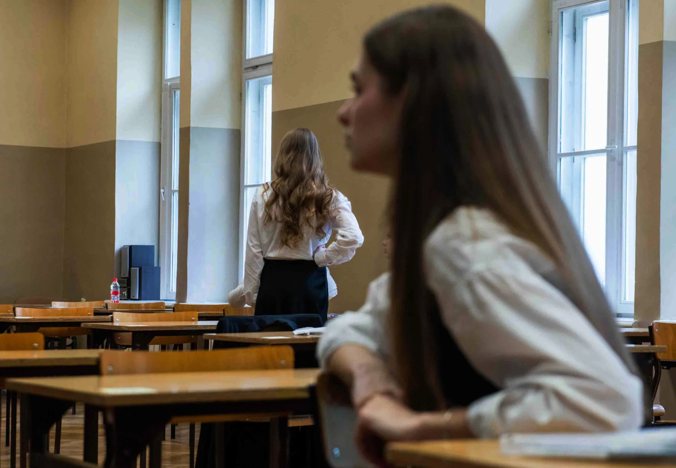 Matura ustna, przecieki. Rodzic alarmuje. "Naruszenie zasad" Uczennice ubrane w białe koszule i ciemne spódnice, jedna z nich siedzi przy ławce, a druga stoi w pobliżu okna, atmosfera klasy szkolnej, drewniane ławki i krzesła, jasne światło wpadające przez wysokie okna.