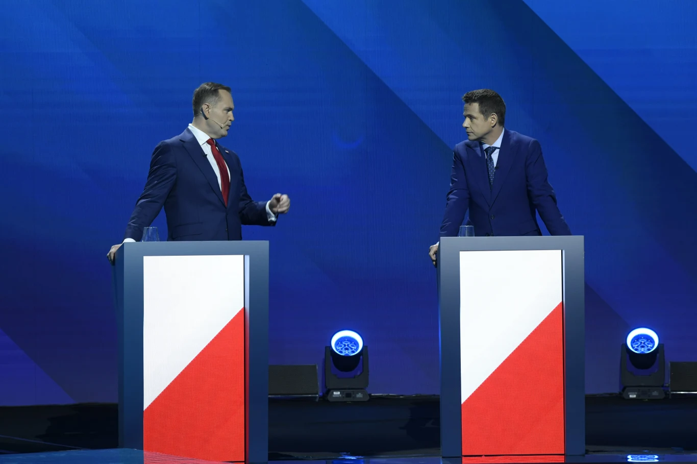 Karol Nawrocki i Rafał Trzaskowski podczas debaty w TVP przed pierwszą turą wyborów prezydenckich 2025 Dwóch mężczyzn w garniturach stoi przy pulpitach ozdobionych biało-czerwonym motywem flagi Polski na tle niebieskiego, nowoczesnego studia telewizyjnego, prowadząc debatę lub dyskusję.