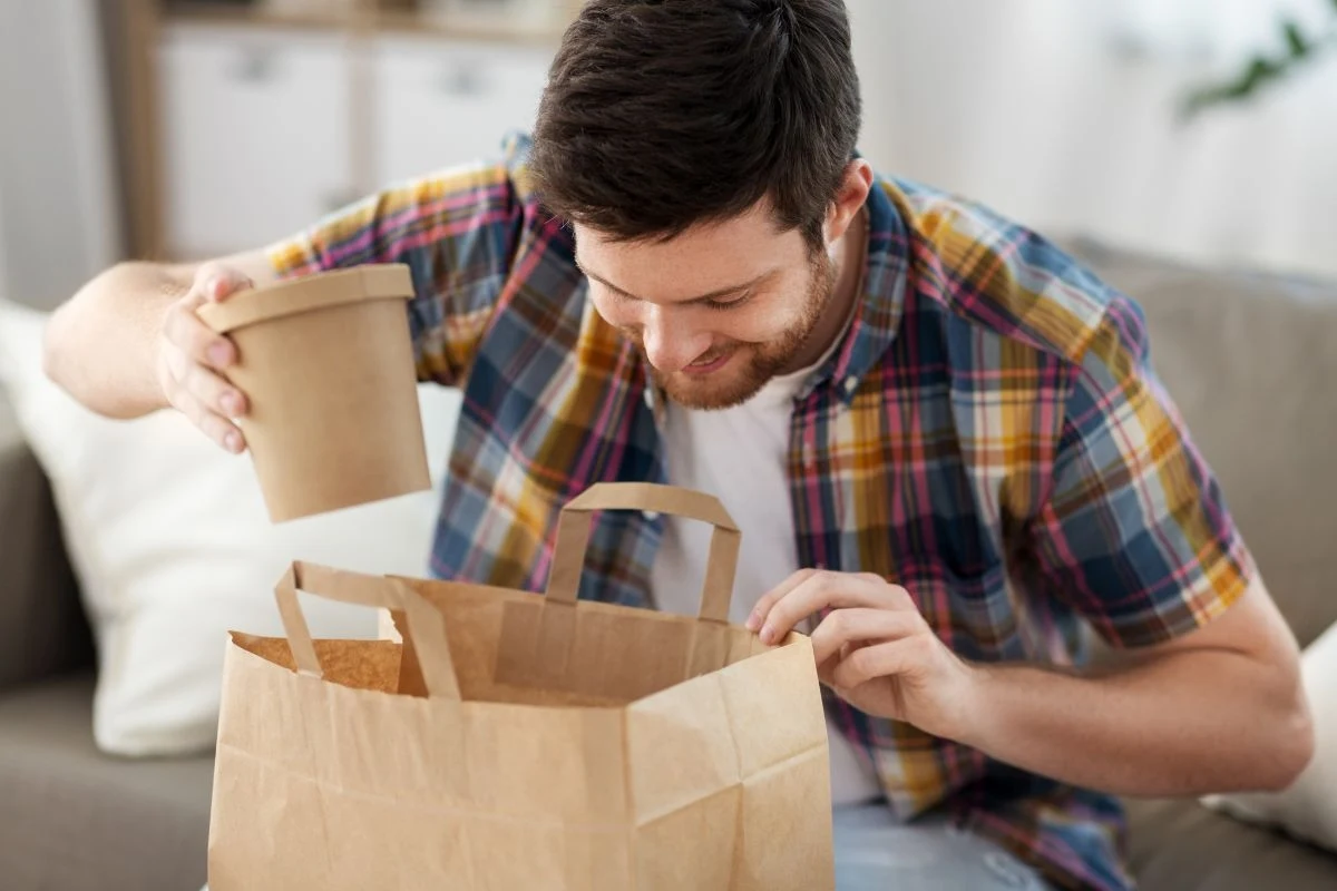 Mężczyzna w koszuli w kratę zagląda do papierowej torby na zakupy, trzymając w ręku papierowy pojemnik z jedzeniem w domowym wnętrzu