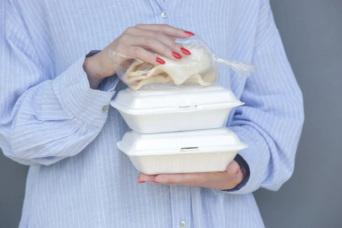 Osoba w jasnej koszuli trzyma dwie białe styropianowe pudełka na jedzenie i plastikowy woreczek z pitą, zwracając uwagę na zadbane paznokcie pomalowane na czerwono.