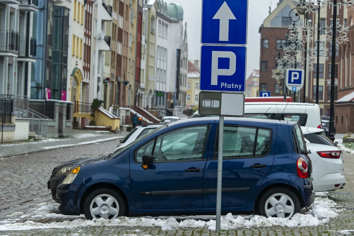 Niebieski samochód zaparkowany na płatnym parkingu miejskim z widocznymi znakami informującymi o strefie parkowania, w tle budynki mieszkalne, fragment ulicy wyłożonej kostką brukową i lekki opad śniegu.
