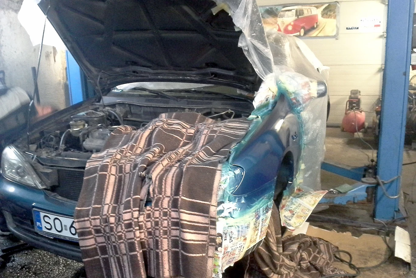 Tak naprawiało się samochody w Polsce jeszcze 10 lat temu / archiwum własne autora Samochód osobowy ustawiony na podnośniku warsztatowym z otwartą maską, częściowo okryty kocem i folią ochronną oraz gazetą, co sugeruje trwające prace naprawcze lub lakiernicze.